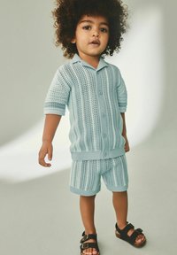 Ensemble en tricot bleu clair comprenant un haut à manches courtes et un short, avec un devant à boutons et un motif texturé. Assorti à des sandales avec boucle noires.