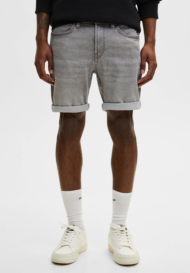 Shorts en jean gris avec ourlets roulés, poches avant et légère décoloration. Portés avec des chaussettes blanches et des baskets de couleur claire. Coupe décontractée.