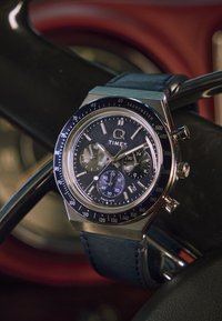 Orologio Timex Q con cassa in acciaio inossidabile, quadrante nero, funzionalità cronografo, sottodial blu e cinturino in pelle blu su un volante.