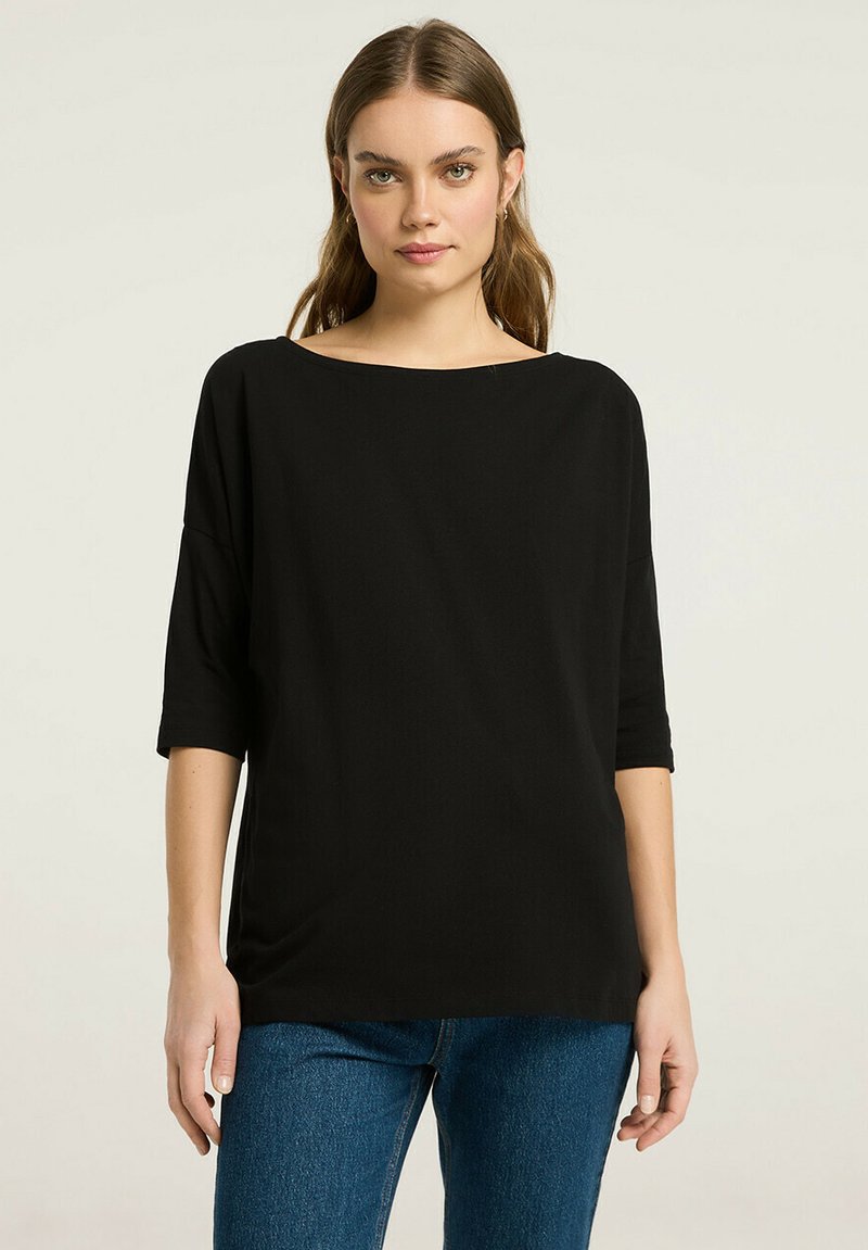 Czarna, luźna bluzka z dekoltem łódki, rękawami do trzech czwartych i miękką fakturą; połączona z niebieskimi dżinsami.