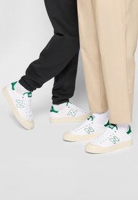 Fehér sneaker zöld részletekkel és texturált krémszínű talppal. Kiemelkedő "N" logóval és lapos fűzőkkel rendelkezik. Sötét és világos nadrággal viselve.