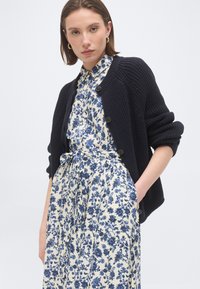 Νεαρή γυναίκα με κοντά καστανά μαλλιά, φοράει χρυσούς κρίκους στα αυτιά, μια σκούρα πλεκτή ζακέτα με κουμπιά πάνω από ένα μπλε και λευκό φόρεμα με floral μοτίβο και ζώνη στη μέση.