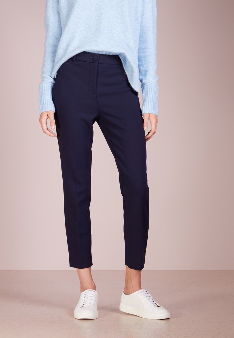 Personne portant un pantalon slim bleu marine aux chevilles, un pull bleu clair et des baskets blanches, debout sur un sol neutre et un fond pâle.
