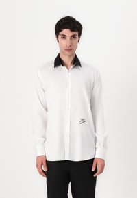 Biela košeľa s button-up zapínaním a čiernym golierom, s dlhými rukávmi a malým vyšitým logom na ľavej strane. Hladká textúra látky.