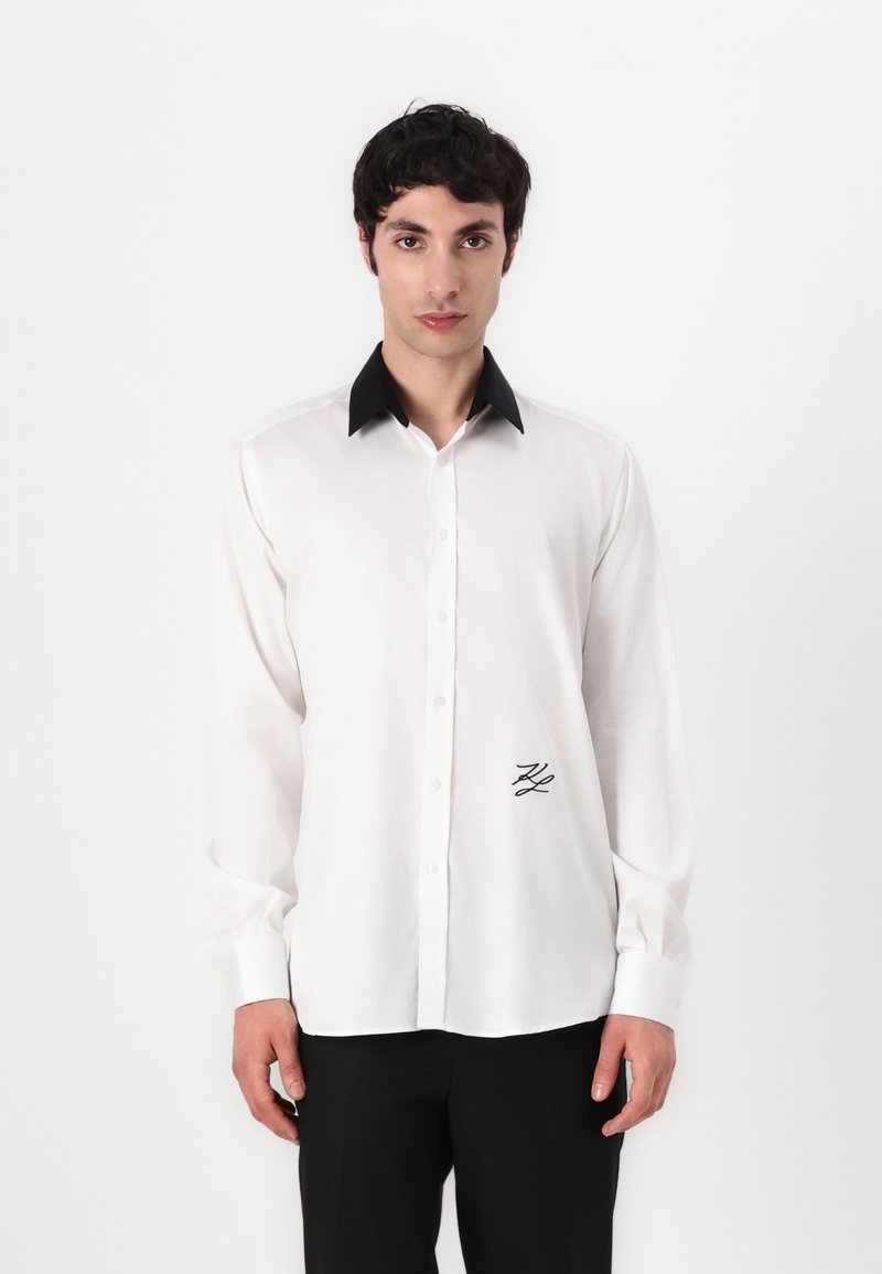 Biela košeľa s button-up zapínaním a čiernym golierom, s dlhými rukávmi a malým vyšitým logom na ľavej strane. Hladká textúra látky.