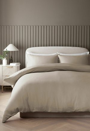 King-Size-Bett mit beiger Bettwäsche und weißen Kissen, einem beigen gepolsterten Kopfteil und einem Beistelltisch mit Lampe und Pflanze.