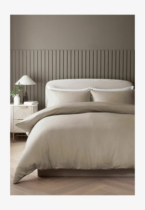 King-Size-Bett mit beiger Bettwäsche und weißen Kissen, einem beigen gepolsterten Kopfteil und einem Beistelltisch mit Lampe und Pflanze.