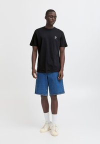 Tricou negru din bumbac cu mâneci scurte și un mic logo pe piept, asortat cu pantaloni scurți din denim albastri, șosete albe și adidași bej.
