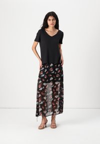 Musthave musta V-kaelusega T-särk, mille juurde on sobitatud durchsichtig maxi seelik, millel on kirglik lehemuster. Modell seisab rihmadega sandaalides ühtlasel taustal.