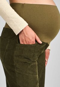 Pantaloni din catifea pentru maternitate, în verde măsliniu închis, cu un braț elastic și neted și buzunare laterale pentru funcționalitate.