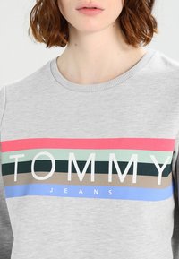 Felpa grigia con scollo rotondo caratterizzata da righe orizzontali multicolori e la scritta "TOMMY JEANS" in bianco sul petto.