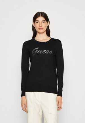 Sort strik sweater med lange ærmer og rund hals. Har et skinnende sølvfarvet "Guess" logo på brystet. Glat tekstur.