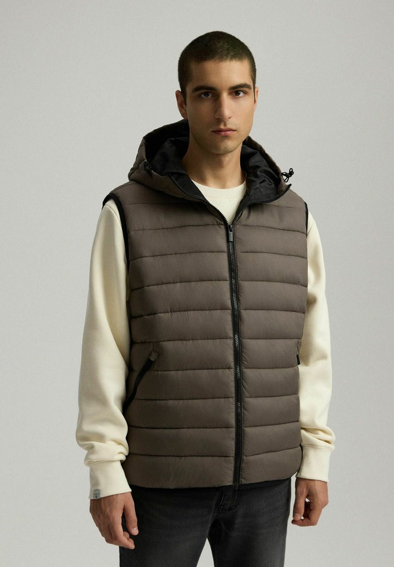 Kamizelka ocieplana w ciemnym brązie z kapturem i czarną podszewką; posiada zamek błyskawiczny i dwie boczne kieszenie. Nosi się ją na jasnej bluzie.