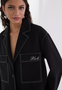 Zwarte blazer met contrasterende witte stiksels. Bevat twee voorzakken, een geknipte revers en een scriptlogo-detail op de zak.