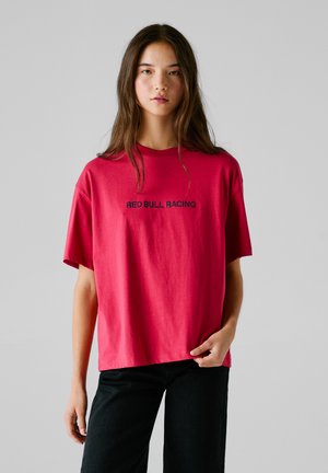 Rotes T-Shirt mit kurzen Ärmeln, mit dem Text "RED BULL RACING" in Schwarz, aus Baumwolle, kombiniert mit schwarzen Hosen.