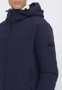 Giacca blu navy con colletto alto, cappuccio regolabile, cerniera frontale e tasche laterali con zip. Presenta una patch con logo sulla manica sinistra.
