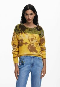 Gelber, gestrickter Pullover mit floralen Mustern in Braun und Grün. Gerippte Bündchen und Ausschnitt. Kombiniert mit hochgeschnittenen blauen, bestickten Jeans.