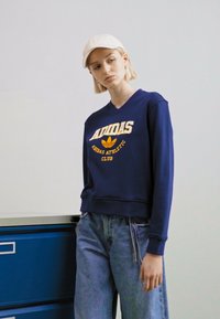 Tamsiai mėlynas džemperis su V formos iškirpte, su geltona "Adidas Athletic Club" tekstu ir logotipu. Dėvimas su šviesiai mėlynomis džinsomis.