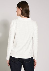 Blusa blanca de manga larga con rayas texturizadas, escote redondeado y un ajuste relajado. Combinada con jeans oscuros en un estilo informal.