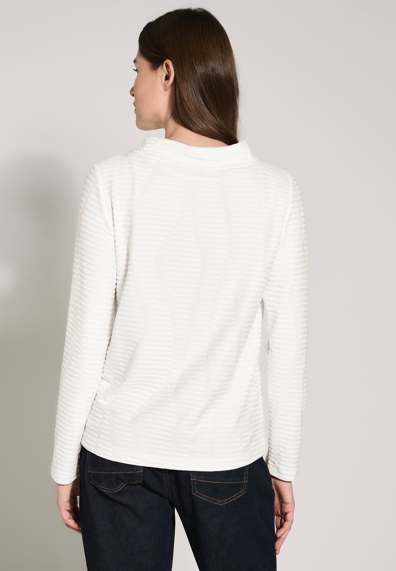 Blusa blanca de manga larga con rayas texturizadas, escote redondeado y un ajuste relajado. Combinada con jeans oscuros en un estilo informal.