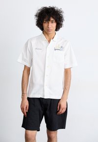 White short-sleeved shirt made of cotton with a pocket. Features embroidered flowers and utensils. Paired with black shorts.