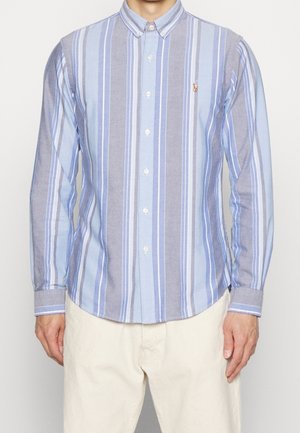 Homme portant une chemise à manches longues boutonnée avec des rayures verticales bleues et grises et un pantalon beige, debout devant un fond uni.