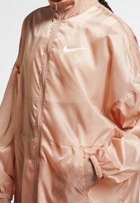 Nike vindjacka i ljus persika, med hög krage, två sidofickor, elastiska ärmmuddar och en distinkt vit logga på bröstet.