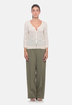 Giovane donna con capelli castani indossa un cardigan bianco trasparente abbottonato e pantaloni larghi verde oliva, in piedi con le braccia rilassate lungo i fianchi.