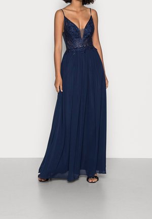 Femme portant une robe de soirée bleue marine sans manches avec un corsage orné de perles, un décolleté en V profond et une jupe fluide en chiffon, debout sur un sol gris.