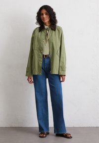 Green lightweight jacket with a brown collar, paired with a ribbed top and wide-leg blue jeans. Features pockets and buttons.