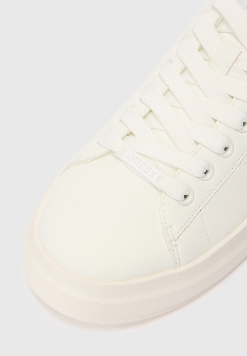 Acercamiento a una zapatilla blanca con cordones y suela de goma, con una etiqueta de cordón de la marca "GUESS" en el material superior liso.