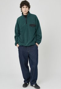 Pull en polaire vert avec un col zippé à mi-hauteur, poche poitrine noire et jean bleu foncé à coupe décontractée. Chaussures noires avec des rayures blanches.