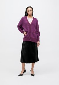 Purple knitted cardigan with long sleeves, button closure, and two front pockets, paired with a black pleated skirt and black heels.