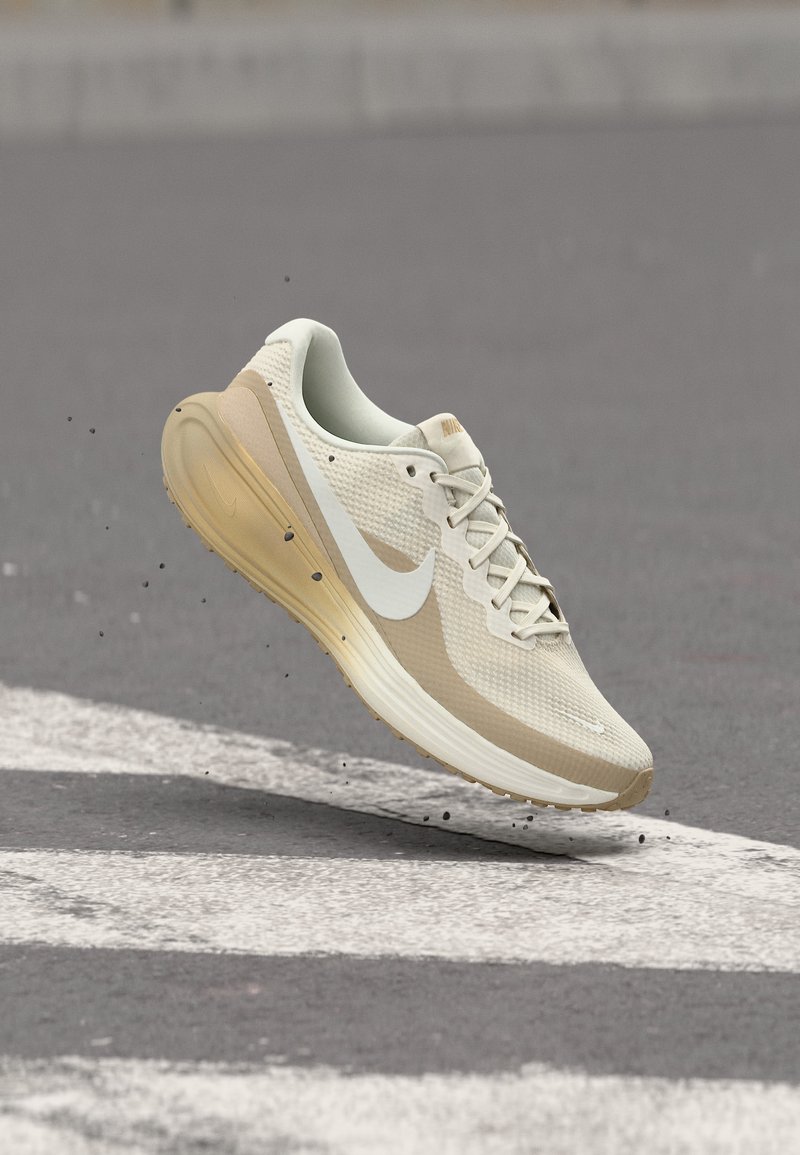 Chaussure de course Nike beige clair et blanche en l'air au-dessus d'une rue avec de la terre éparpillée sur l'asphalte gris.