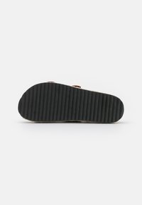 Zwarte rubberen zool met horizontale ribbels, contrasterende bruine bovenkant met geweven textuur en twee riemaccenten.
