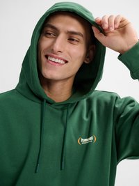 Junger Mann lächelt, trägt einen grünen Hoodie mit kleinem "hummel SPORTSWEAR"-Logo und hebt die Kapuze mit der rechten Hand vor schlichtem Hintergrund.