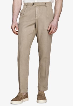 Pantalones beige a medida con puños, combinados con mocasines de ante marrón, en una persona de pie que lleva una camisa blanca.