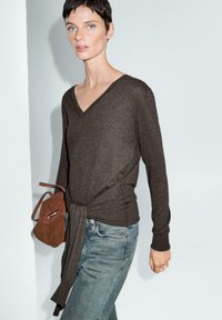 Brauner V-Ausschnitt-Pullover mit Wickeldetail, kombiniert mit hellblauen Jeans. Das Model hält eine kleine braune Wildledertasche. Glatte Textur, moderner Schnitt.