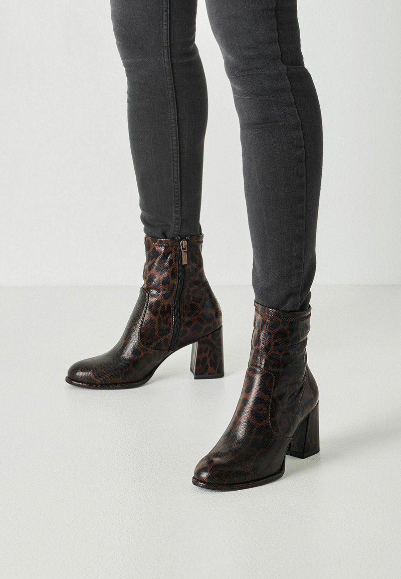 Personne portant des bottines foncées à imprimé léopard avec talons blocs et un jean skinny noir, debout sur un sol clair.