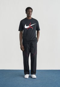 Μαύρο κοντομάνικο μπλουζάκι με λευκό και κόκκινο λογότυπο Nike. Μαύρο παντελόνι με κόκκινες λεπτομέρειες. Λευκά αθλητικά παπούτσια. Ουδέτερο φόντο.