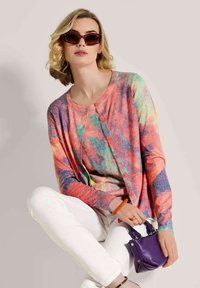 Frau in einem bunten, geknöpften Cardigan, weißen Hosen, dunkell roten Sonnenbrille, die eine kleine lila Handtasche hält, während sie im Schneidersitz sitzt.