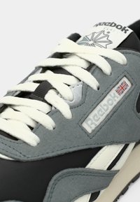 Gros plan d’une basket Reebok Classic grise et noire avec des lacets blancs et un drapeau Union Jack sur le patch du logo.