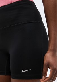 Κοντινό πλάνο ατόμου που φοράει μαύρα αθλητικά σορτς της Nike με ασημένιο λογότυπο Swoosh στον αριστερό μηρό, και ένα ροζ τοπ που φαίνεται εν μέρει.