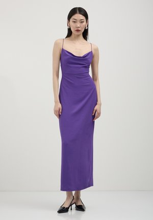 Una donna in un abito lungo senza maniche color viola con spalline sottili e tacchi a punta neri, in piedi contro uno sfondo grigio chiaro uniforme.