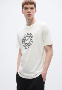 Balti medvilniniai marškinėliai su apvalia iškirpte ir trumpomis rankovėmis, puošti juodos spalvos apvaliu grafiniu dizainu, kuriame yra tekstas ir teniso kamuoliuko motyvas.