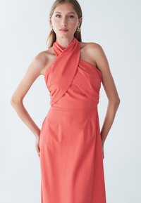 Korallpunane halter-kaelusega kleit, millel on mähitud kangadisain, keha järgiv siluett ja sile tekstuur. Puuduvad nähtavad mustrid või metalldetailid.