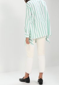 Žena v bílých kalhotách s roztrhanými lemy, černých nazouvacích botách a volné bílé košili se zelenými vertikálními pruhy stojící před jednobarevným pozadím.