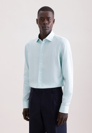 Homme debout, de face, portant une chemise rayée bleu clair à manches longues boutonnée et un pantalon bleu marine foncé sur un fond uni.