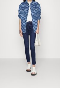 Chaqueta vaquera con patrón geométrico en azul, camiseta blanca, vaqueros ajustados oscuros y sandalias blancas de punta abierta con suelas negras. Look casual.