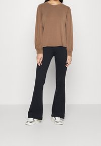 Maglietta marrone a maniche lunghe con spalle a palloncino, abbinata a pantaloni aderenti neri a zampa d'elefante e sneakers metallizzate argento. Design semplice e casual.